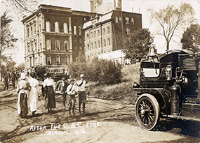 Three young boys walk away while three women walk toward the shell of the Christian Brothers College buildings that had been severely damaged in an early morning fire.  A fire truck is parked on the site.  Firemen and police are also in the group of onlookers.